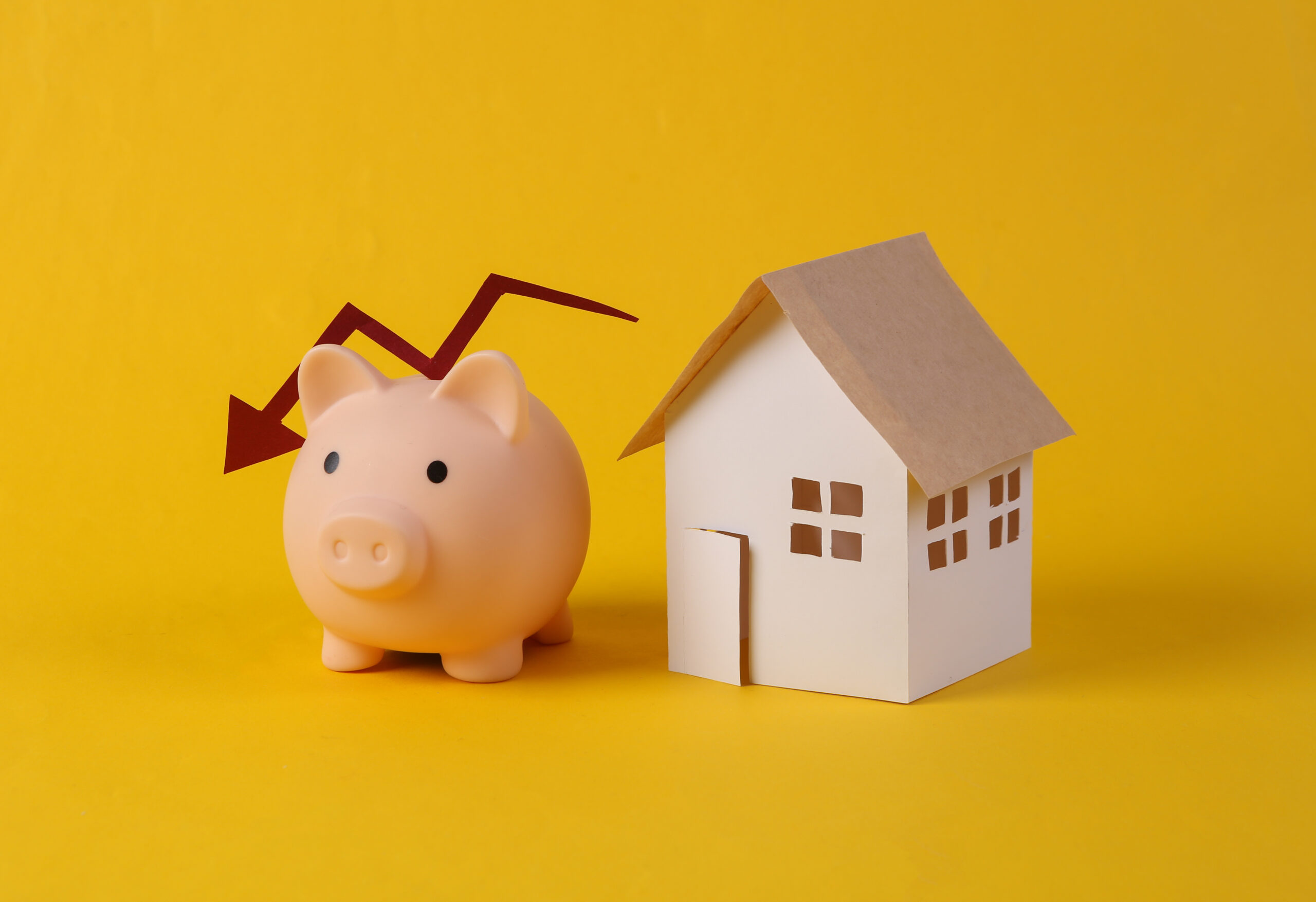 Falling housing prices. House figurine and piggy bank with falling arrow on yellow background
