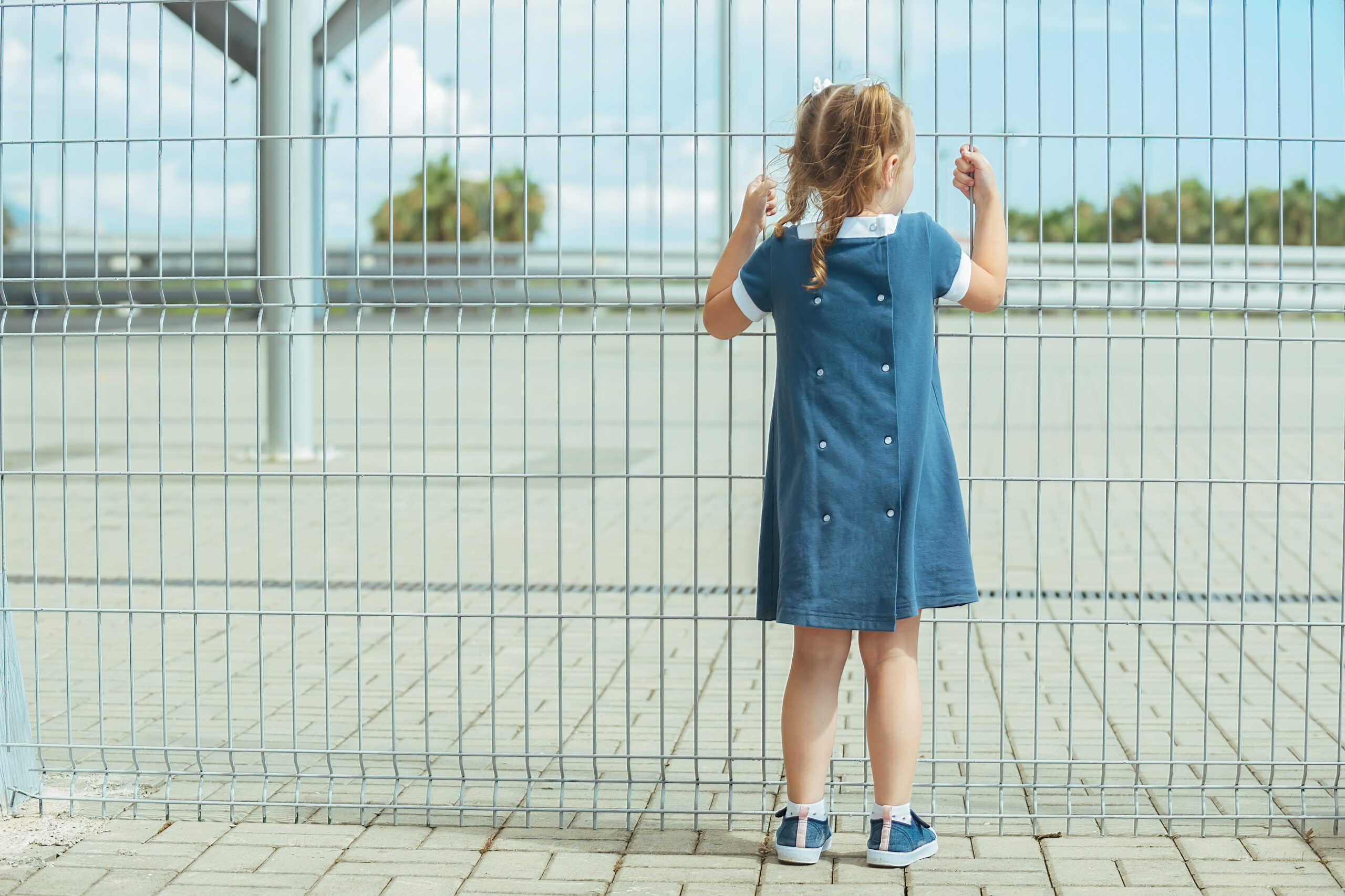 preschool-girl-in-blue-dress-stands-near-barrier-from-high-mesh-fence-and-holds-on-to-it
